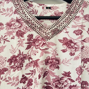 May include: A white top with a V-neckline, adorned with a floral pattern in shades of burgundy and pink. The neckline is embellished with intricate embroidery. The fabric is draped over a black hanger.