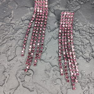Pode incluir: Um par de brincos pendurados com uma parte superior retangular e vários fios de pequenas pedras rosa. Os brincos estão sobre um fundo escuro e texturizado.