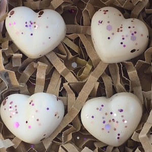 Op de afbeelding: Vier hartvormige waxmelts in een gebroken witte kleur, elk versierd met verspreide zeshoekige en ronde glitters in roze, paarse en koperen tinten, gerangschikt in een vierkante formatie in een doos gevuld met bruine papiersnippers.