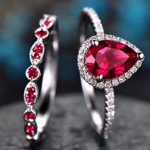 Puede incluir: Un conjunto de dos anillos que incluye una sortija con una piedra roja en forma de pera rodeada por un halo de pequeñas piedras blancas sobre una banda plateada, junto a una alianza a juego con pequeñas piedras rojas y blancas alternadas.