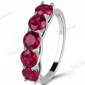 Può includere: Un anello color argento con cinque rubini rosso scuro a taglio rotondo disposti in una fila verticale lungo la fascia. La fascia metallica ha una finitura lucida e le pietre sono fissate con griffe, creando una linea continua di gemme rosse.