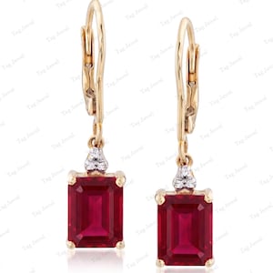 Pode incluir: Um par de brincos pendentes com gemas vermelhas de corte esmeralda retangular cravadas em ouro amarelo. Cada brinco possui um fecho de alavanca e um pequeno grupo de três minúsculas pedras brancas acima da gema vermelha principal.