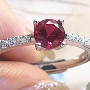 Könnte beinhalten: Ein silberfarbener Ring mit einem runden, tiefroten Mittelstein, der von vier Krappen gehalten wird. Die Ringschiene ist auf beiden Seiten des zentralen Steins mit einer Reihe kleiner, klarer, funkelnder Steine besetzt.