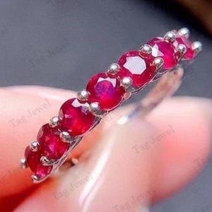 May include: A silver-toned band ring featuring a row of seven round-cut, vibrant red rubies set in a prong setting. The gemstones are arranged in a straight line along the top of the metal band, showcasing a polished finish and deep red colour.