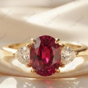 Op de afbeelding: Een gouden ring met een grote, ovaal geslepen, dieprode edelsteen in het midden, geflankeerd door twee kleinere, heldere, driehoekig geslepen stenen aan weerszijden. De ring is gezet op een gladde, gepolijste gouden band met een klassieke zetting.