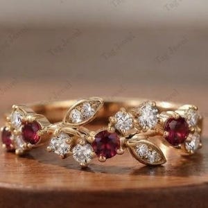 Op de afbeelding: Een gouden ring met een rankontwerp, voorzien van afwisselend ronde rode edelstenen en heldere witte stenen. De band is afgewerkt met metalen blaadjes tussen de stenen, wat zorgt voor een doorlopend bloemmotief rondom de ring.