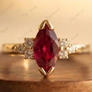Peut inclure: Une bague en or avec une pierre précieuse rouge foncé taille marquise fixée par des griffes. L'anneau est orné de chaque côté d'un groupe de trois petites pierres blanches rondes et claires, formant un design symétrique en or jaune poli.
