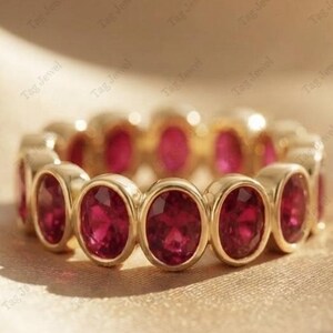 Könnte beinhalten: Ein goldener Eternity-Ring mit einer durchgehenden Reihe von oval geschliffenen, tiefroten Rubinen, die in einzelnen polierten Goldfassungen sitzen. Der Ring zeigt ein einheitliches Design, bei dem jeder Edelstein in Metall gefasst ist.