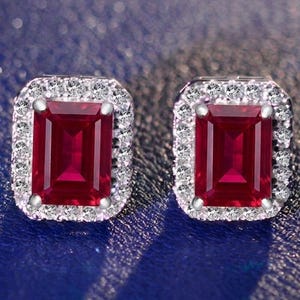 May include: A pair of stud earrings featuring rectangular emerald-cut red gemstones set in silver-toned metal. Each central red stone is surrounded by a halo of small, clear, round-cut white stones, creating a bright and reflective appearance.