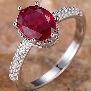 Op de afbeelding: Een zilverkleurige ring met een grote, ovaal geslepen dieprode edelsteen in een zetting met zes pootjes. De ringband is aan de zijkanten bezet met kleine, heldere pavé-stenen, wat zorgt voor een sprankelend effect op het metaal.