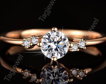 May include: A rose gold ring featuring a central round brilliant-cut clear stone flanked by two smaller clear stones on each side. The band is smooth and polished, reflecting light against a dark background with the repeated text Tagjewel.
