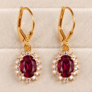 May include: A pair of gold leverback earrings featuring oval-cut deep red rubies surrounded by a halo of small, clear, round-cut white stones. The earrings are displayed symmetrically, showcasing the vibrant red gems and the polished gold metal.