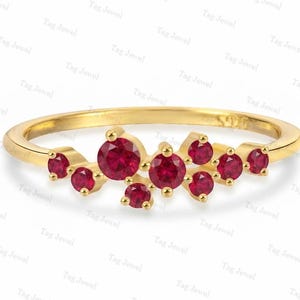 Può includere: Un anello in oro con un gruppo di nove pietre preziose rosse a taglio rotondo disposte in un motivo asimmetrico e sfalsato sulla parte superiore della fascia. La fascia in metallo è liscia e lucida, con una tonalità oro giallo brillante.