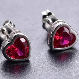 Puede incluir: Un par de pendientes de botón con piedras rojas facetadas en forma de corazón engastadas en metal plateado pulido. Cada pendiente incluye un cierre de presión seguro, mostrando un tono carmesí vibrante y un diseño de silueta de corazón.