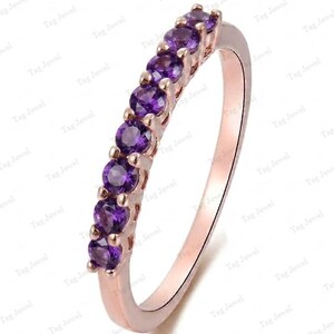 Op de afbeelding: Een roségouden ring met een rij van zeven rondgeslepen paarse amethist edelstenen in een klauwzetting. De metalen band heeft een gladde, gepolijste afwerking en de stenen zijn in een rechte lijn langs de bovenste helft van de ring geplaatst.