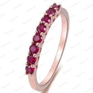 Könnte beinhalten: Ein Ring aus Roségold mit einer Reihe von sieben rund geschliffenen, tiefroten Rubinen in einer gemeinsamen Krappenfassung. Das Metall hat eine polierte Oberfläche und die Steine sind in einer geraden Linie auf der oberen Ringhälfte angeordnet.