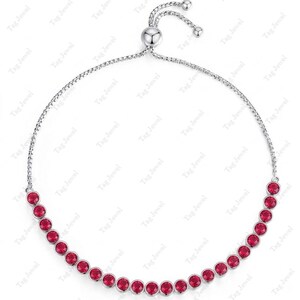 Op de afbeelding: Een verstelbare bolo-armband in zilverkleur met een rij ronde, felrode edelstenen gezet in individuele metalen zettingen. De armband is voorzien van een schuifmechanisme met twee bungelende kettinguiteinden voor een aanpasbare pasvorm.