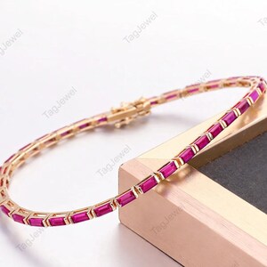 Könnte beinhalten: Ein goldfarbenes Tennisarmband mit einer durchgehenden Reihe rechteckiger, im Baguetteschliff geschliffener, leuchtend pinker Edelsteine in einer Metallfassung. Das Armband verfügt über einen sicheren Kastenschloss-Verschluss und liegt auf einer goldgeränderten Oberfläche.