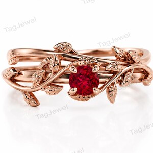 May include: A rose gold ring featuring a central round-cut red gemstone. The band is designed with intricate, overlapping vine and leaf motifs that wrap around the finger, creating a nature-inspired aesthetic with a polished metallic finish.
