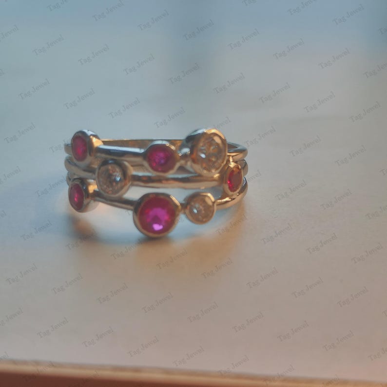 Op de afbeelding: Een zilveren ring met meerdere banden en een reeks ronde stenen in een zetting. Het ontwerp bevat afwisselend felroze robijnen en heldere witte stenen die in een asymmetrisch patroon over de ringbanden zijn verdeeld.