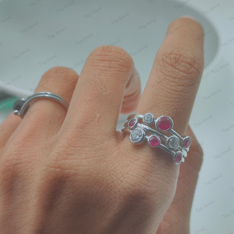 Op de afbeelding: Een zilverkleurige ring met meerdere banden en een cluster van in een zetting geplaatste ronde stenen, afwisselend heldere witte edelstenen en levendige rode robijnen. Het ontwerp bestaat uit verschillende dunne, met elkaar verbonden banden.