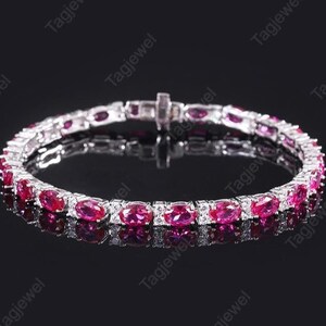 Puede incluir: Una pulsera de plata con piedras preciosas rojas vibrantes de corte ovalado y piedras más pequeñas y transparentes. La pulsera se muestra sobre una superficie oscura y reflectante, resaltando el brillo de las gemas. El cierre es visible y el diseño es elegante.