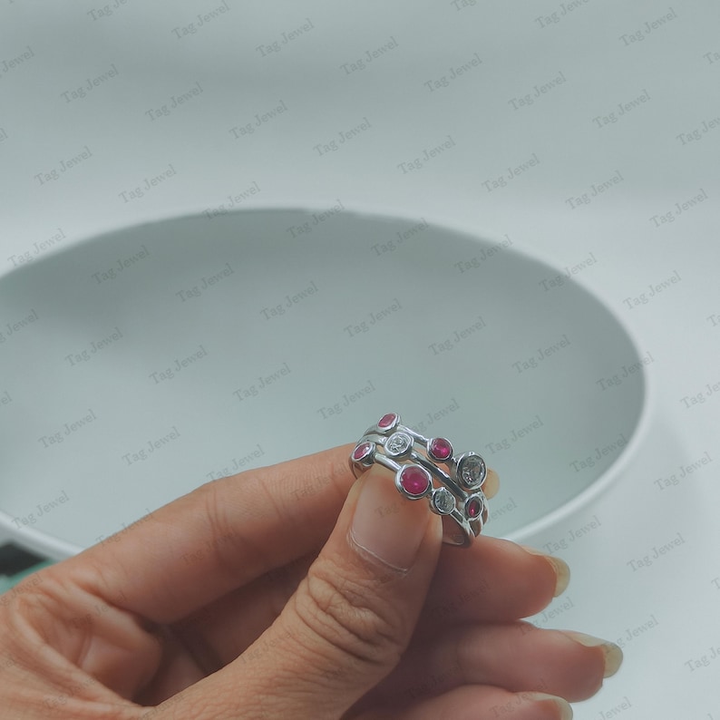 Op de afbeelding: Een zilverkleurige ring met een ontwerp van meerdere banden en rond gezette stenen. De stenen wisselen af tussen helderwitte en felroze kleuren, wat zorgt voor een gelaagd uiterlijk over het oppervlak van de metalen bandstructuur.