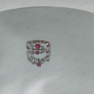 Op de afbeelding: Een zilverkleurige ring met een ontwerp van meerdere banden en in chaton gezette ronde stenen. De ring is versierd met afwisselend felrode ronde edelstenen en heldere, kleurloze ronde stenen in een asymmetrisch patroon.