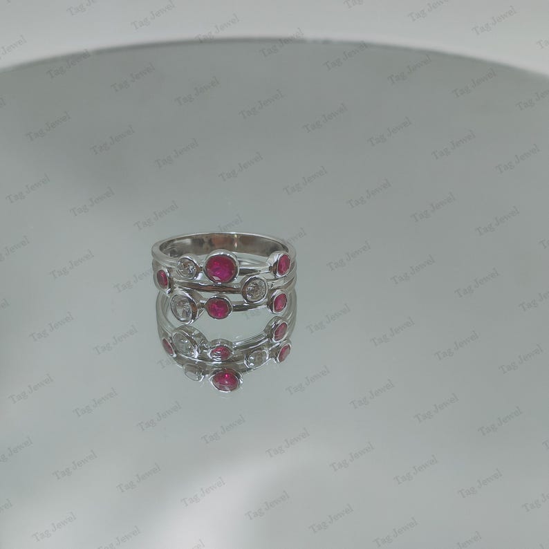 Op de afbeelding: Een zilverkleurige ring met een ontwerp van meerdere banden en in chaton gezette ronde stenen. De ring is versierd met afwisselend felrode ronde edelstenen en heldere, kleurloze ronde stenen in een asymmetrisch patroon.