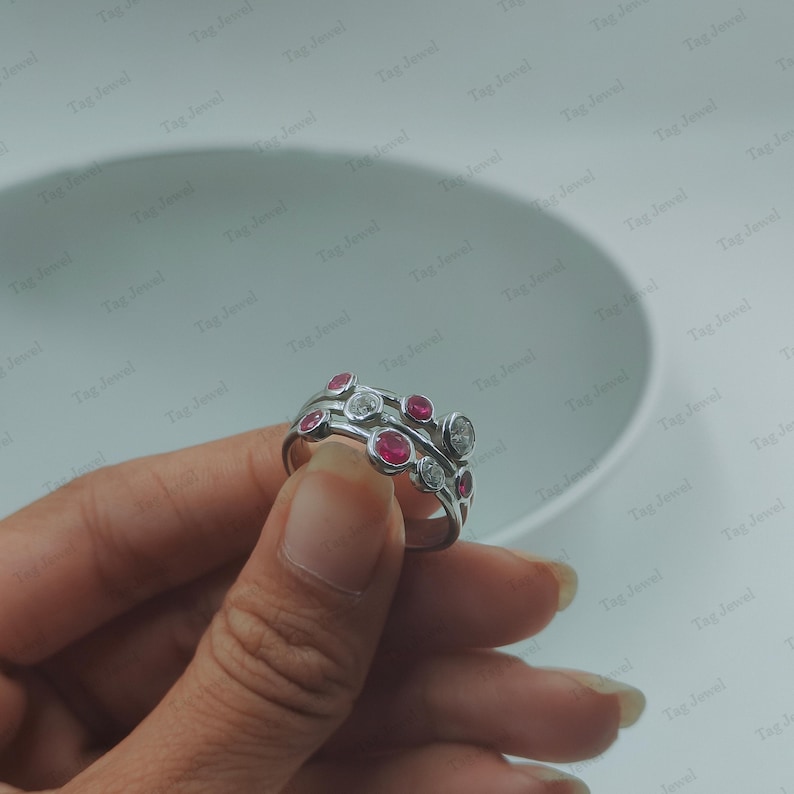 Op de afbeelding: Een zilverkleurige ring met een ontwerp van meerdere banden en in een zetting geplaatste ronde stenen. De ring is versierd met afwisselend felrode ronde stenen en heldere, kleurloze ronde stenen, allemaal vastgezet in een gepolijste metalen zetting.