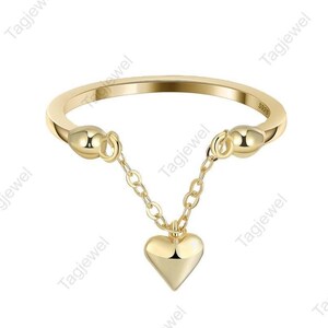 May include: A gold-colored ring with a heart pendant. The ring features a delicate chain that suspends a polished heart charm. The ring is a simple band with decorative accents on each side of the chain.