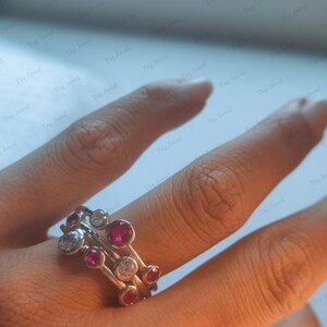 Op de afbeelding: Een zilveren ring met een ontwerp van meerdere banden en in een zetting geplaatste ronde stenen. De ring is versierd met afwisselend heldere witte stenen en levendige rode stenen, wat een asymmetrisch patroon op de metalen banden vormt.