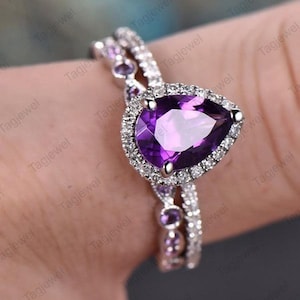 May include: A silver ring set featuring a pear-shaped, deep purple gemstone surrounded by small, clear stones. The band is also adorned with smaller purple gemstones. The ring is designed to be worn as a set.