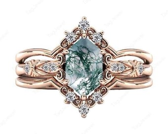 May include: A rose gold ring set featuring an ornate design. The engagement ring has a central, emerald-cut, moss agate stone, surrounded by small, clear gemstones. The band is accompanied by two additional bands with similar detailing.