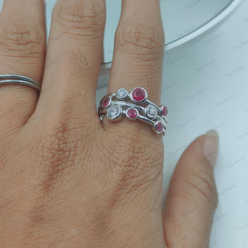 Op de afbeelding: Een zilverkleurige ring met een ontwerp van meerdere banden, voorzien van ronde rode stenen en heldere witte stenen in een chatonzetting, asymmetrisch gerangschikt over het oppervlak van de ring, gedragen naast een eenvoudige zilverkleurige band.
