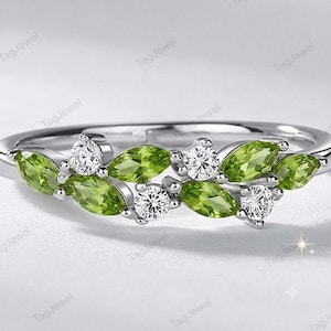 May include: A silver-toned ring featuring a cluster of marquise-cut green peridot stones and round-cut clear white stones arranged in a leaf-like pattern along the band. The metal band is polished and smooth, showcasing a delicate botanical design.