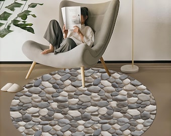 May include: A round area rug with a pebble stone design in shades of gray and white. The rug is placed on a brown floor, with a gray armchair and a floor lamp in the background. The rug adds a natural, textured element to the room.