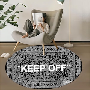 以下が含まれることがあります： 白と黒の模様が入った楕円形のラグで、白い文字で「KEEP OFF」と書かれています。グレーのアームチェアに座って本を読んでいる人がいます。ラグは茶色の床にあります。