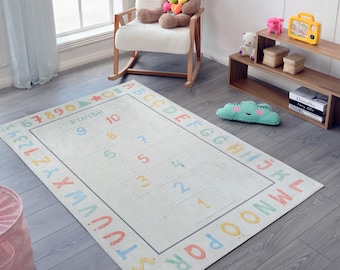 May include: A rectangular off-white hopscotch rug with a colorful border of letters and numbers. The hopscotch grid is in the center, with numbers 1-10. The rug is on a gray wooden floor.