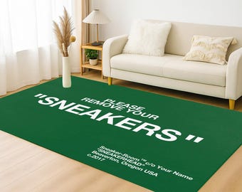 May include: Green rectangular rug with the text "PLEASE REMOVE YOUR SNEAKERS" in white. The rug also includes the text "Sneaker-Room" and the location "Beaverton, Oregon USA". A white sofa and a vase with dried plants are in the background.