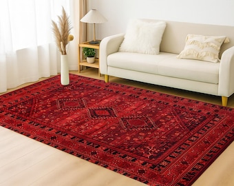May include: A large, rectangular red rug with an intricate geometric pattern. The rug features a central diamond design and a detailed border. It is placed on a light-colored wooden floor, with a white sofa and decorative items in the background.