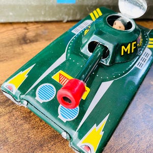 May include: A vintage green toy tank with yellow and white accents, a red barrel, and the letters "MF" in yellow. A small figure is visible in the turret. The toy is on a wooden surface.