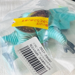 May include: A blue and brown toy figure of a creature, packaged in a clear plastic bag. The figure has a textured head and a segmented body. A yellow tag with Japanese text is attached to the bag, along with a white label with a barcode and Japanese text.