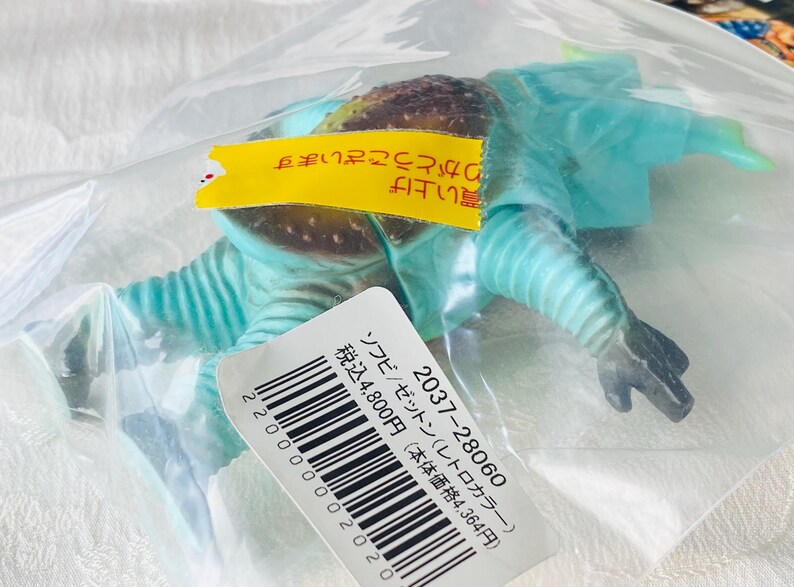 May include: A blue and brown toy figure of a creature, packaged in a clear plastic bag. The figure has a textured head and a segmented body. A yellow tag with Japanese text is attached to the bag, along with a white label with a barcode and Japanese text.
