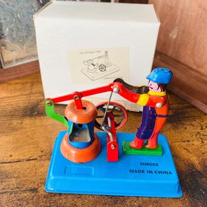 以下が含まれることがあります： 青い帽子とオレンジ色のジャケットを着た人物が、赤と緑の機械を操作するカラフルなヴィンテージブリキのおもちゃ。おもちゃは青いベースにあり、「MADE IN CHINA」と印刷されています。背景には白い箱があります。