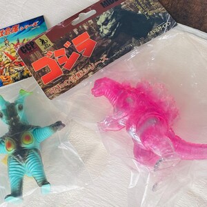 May include: A clear plastic bag contains two toy figures and packaging. One figure is a translucent pink Godzilla. The other is a blue and green monster with outstretched arms. Packaging includes Japanese text and the word "Godzilla".