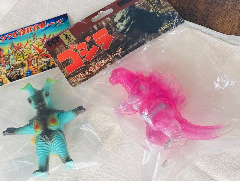 May include: A clear plastic bag contains two toy figures and packaging. One figure is a translucent pink Godzilla. The other is a blue and green monster with outstretched arms. Packaging includes Japanese text and the word "Godzilla".