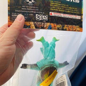 May include: A teal and brown vinyl figure of a monster in a clear plastic bag. The figure has outstretched arms and green head appendages. The packaging includes Japanese text and a warning label stating "Not a Child's Toy."