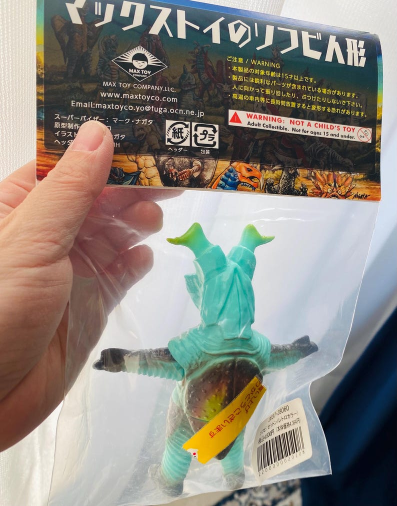 May include: A teal and brown vinyl figure of a monster in a clear plastic bag. The figure has outstretched arms and green head appendages. The packaging includes Japanese text and a warning label stating "Not a Child's Toy."