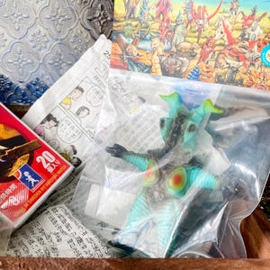 May include: A teal and black monster action figure in a plastic bag, with red and yellow accents. A box of matches with Japanese text and a comic strip are also visible.